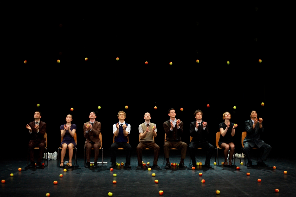 9 comédiens assis sur scène face au public avec des pommes à leurs pieds