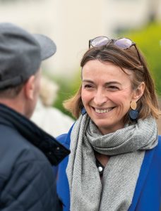 Une femme souriante, portant des lunettes de soleil et un foulard, est photographiée en extérieur.