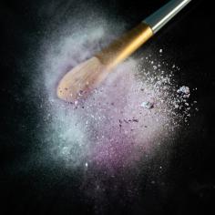Sur un fond noir pur, un pinceau de maquillage, chargé de poudre de pigments vibrants, est soumis à des vibrations intenses.