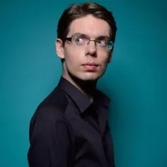 Un homme brun avec des lunettes pose devant l'objectif avec un fond bleu turquoise