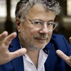 Un homme avec des cheveux grisonnant et des lunettes fixe la caméra