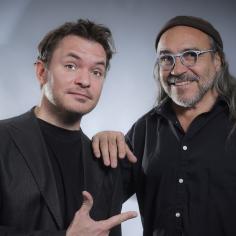 Deux hommes dont un avec des cheveux long et des lunettes sourient à la caméra