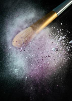Sur un fond noir pur, un pinceau de maquillage, chargé de poudre de pigments vibrants, est soumis à des vibrations intenses.