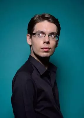 Un homme brun avec des lunettes pose devant l'objectif avec un fond bleu turquoise