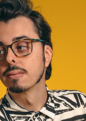 Un homme brun avec des lunettes regarde à droite et le fond est jaune-orangé