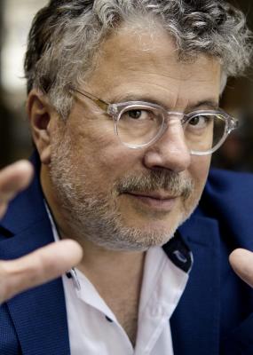 Un homme avec des cheveux grisonnant et des lunettes fixe la caméra