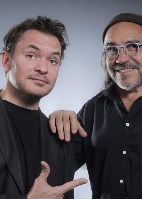 Deux hommes dont un avec des cheveux long et des lunettes sourient à la caméra