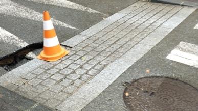 Un convoi orange et un passant piéton sur une chaussée avec des travaux et un égout.