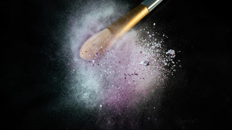 Sur un fond noir pur, un pinceau de maquillage, chargé de poudre de pigments vibrants, est soumis à des vibrations intenses.