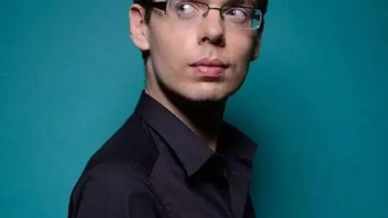 Un homme brun avec des lunettes pose devant l'objectif avec un fond bleu turquoise