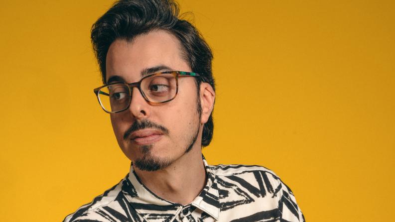Un homme brun avec des lunettes regarde à droite et le fond est jaune-orangé
