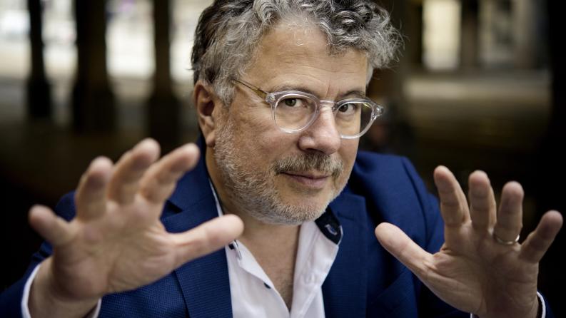 Un homme avec des cheveux grisonnant et des lunettes fixe la caméra