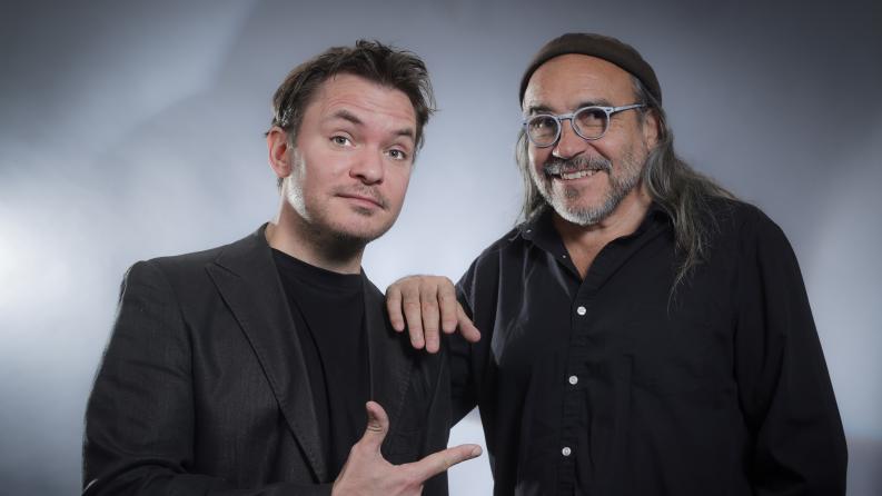 Deux hommes dont un avec des cheveux long et des lunettes sourient à la caméra