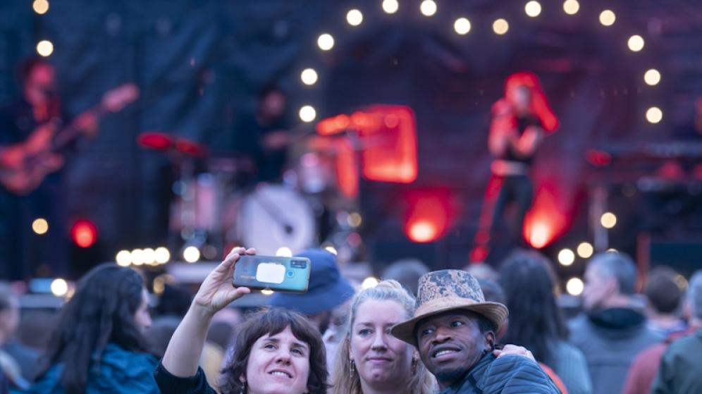 On voit trois personnes qui prennent une photo et dans le fond on aperçoit une scène de concert