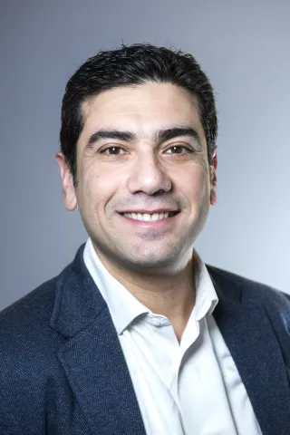 Un homme brun portant une chemise blanche et une veste bleu sourit à l'écran