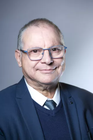 Un homme âgé aux cheveux gris et lunettes bleus sourit à l'écran