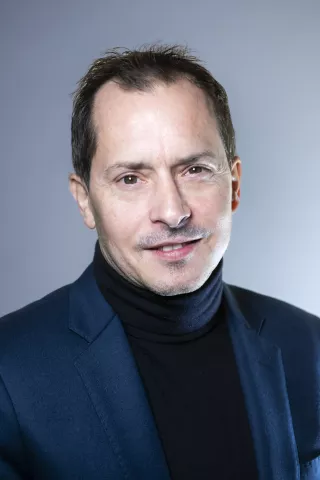 Un homme brun avec un col roulé sourit à l'écran