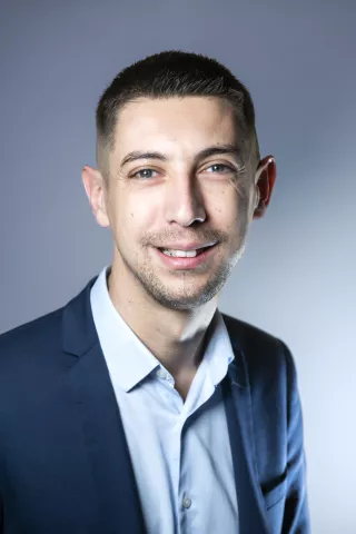 Un homme brun portant une chemise bleu sourit à l'écran