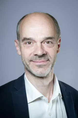 Un homme chauve avec une chemise blanche et une veste noire sourit à l'écran