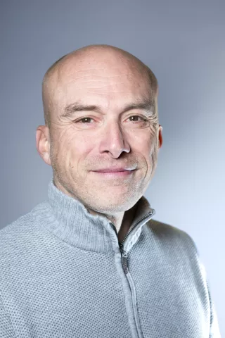 Un homme chauve avec un gilet gris sourit à l'objectif 